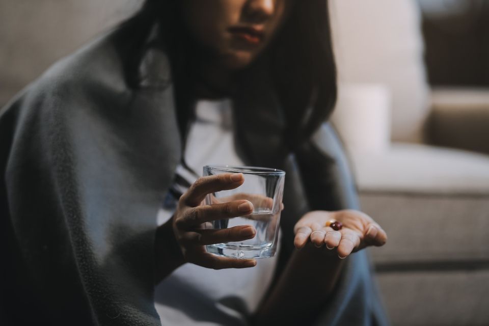 Woman with blanket around her shoulders, holding pills in one hand and water in the other.