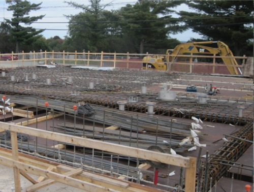 Chantier de construction avec barres d'armature, coffrages en béton et excavatrice ; une clôture en bois entoure la zone.