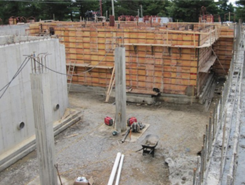 Chantier : Murs en béton et coffrages en cours de réalisation. Charpente en bois aux tons naturels, armatures apparentes et outils sont visibles.