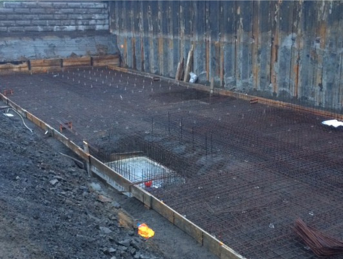 Chantier avec treillis d'armature et coffrage en bois. Un mur métallique et une ouverture carrée sont visibles.