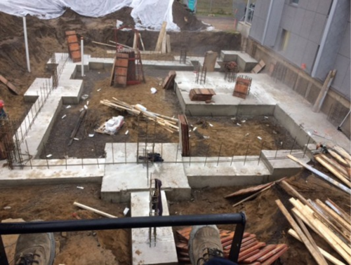 Chantier de construction avec fondations en béton et armatures apparentes, entouré de terre et de planches de bois.