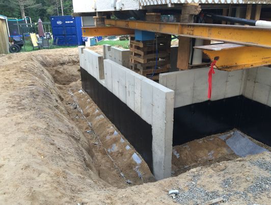 Fondations en béton en construction, la maison étant surélevée au-dessus, laissant apparaître la membrane d'étanchéité.
