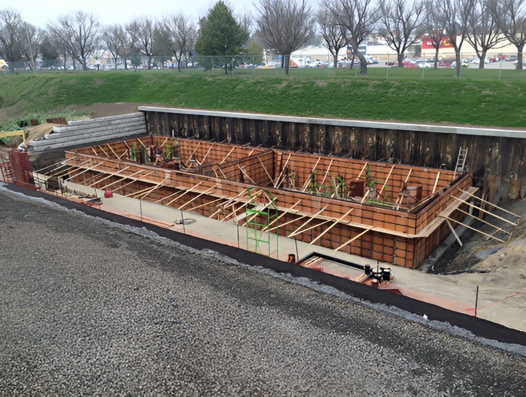 Chantier : Coffrages en bois pour une structure en béton, construite à flanc de colline, avec murs de soutènement et route au premier plan.