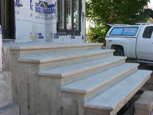 Des marches en béton mènent à l'entrée d'un bâtiment. Les marches sont arrondies et de couleur gris clair. Un camion blanc est stationné à proximité.