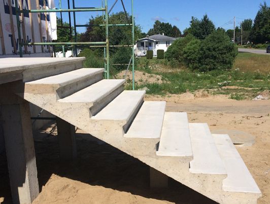 Escalier en béton en construction menant à un bâtiment ; environnement extérieur avec sol sablonneux.