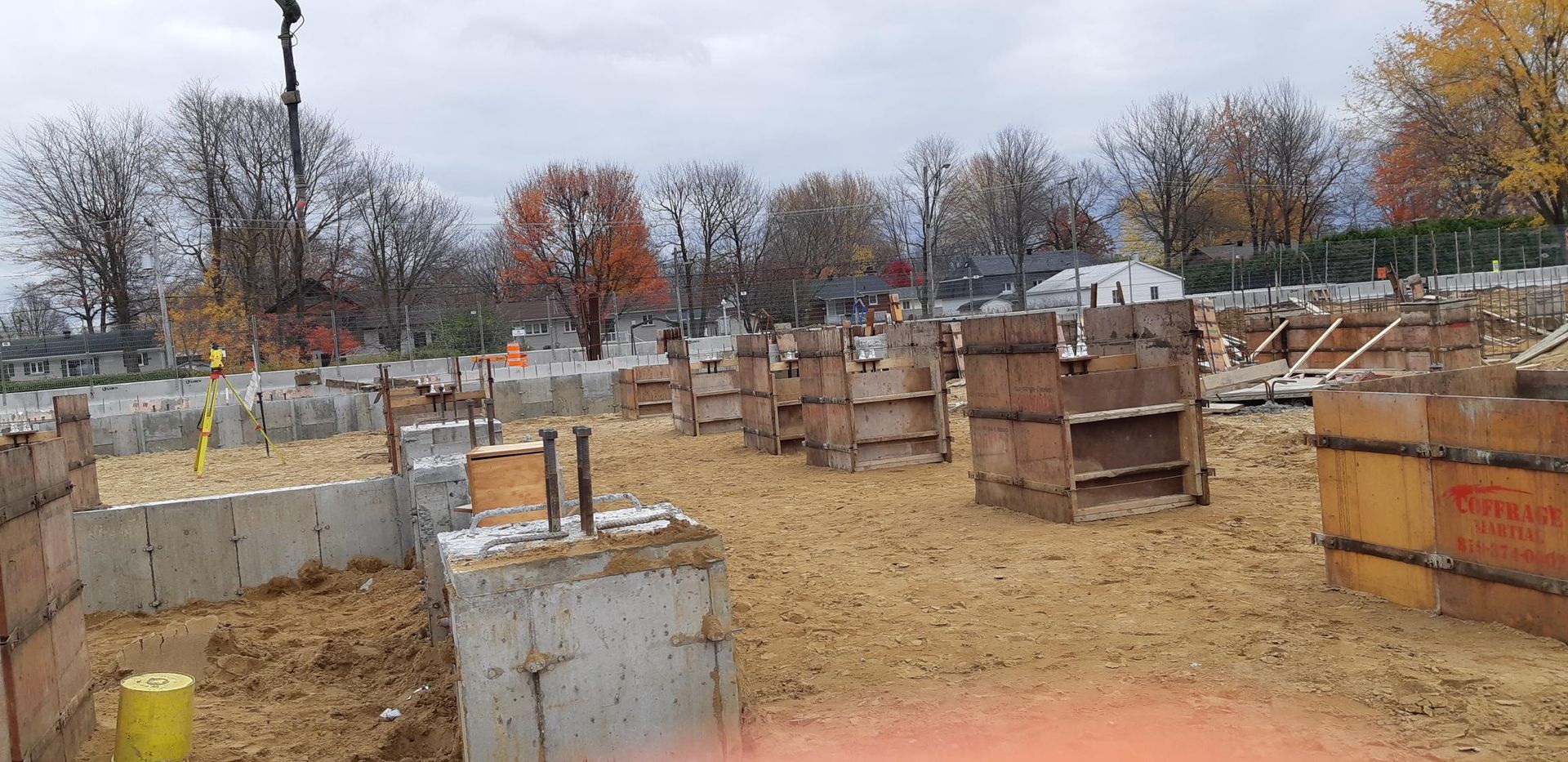 Chantier de construction avec fondations en béton et coffrages en bois, ciel nuageux et arbres d'automne en arrière-plan.