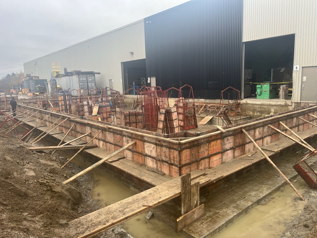 Chantier : Coffrages en béton et armatures devant un grand bâtiment à travées ouvertes.