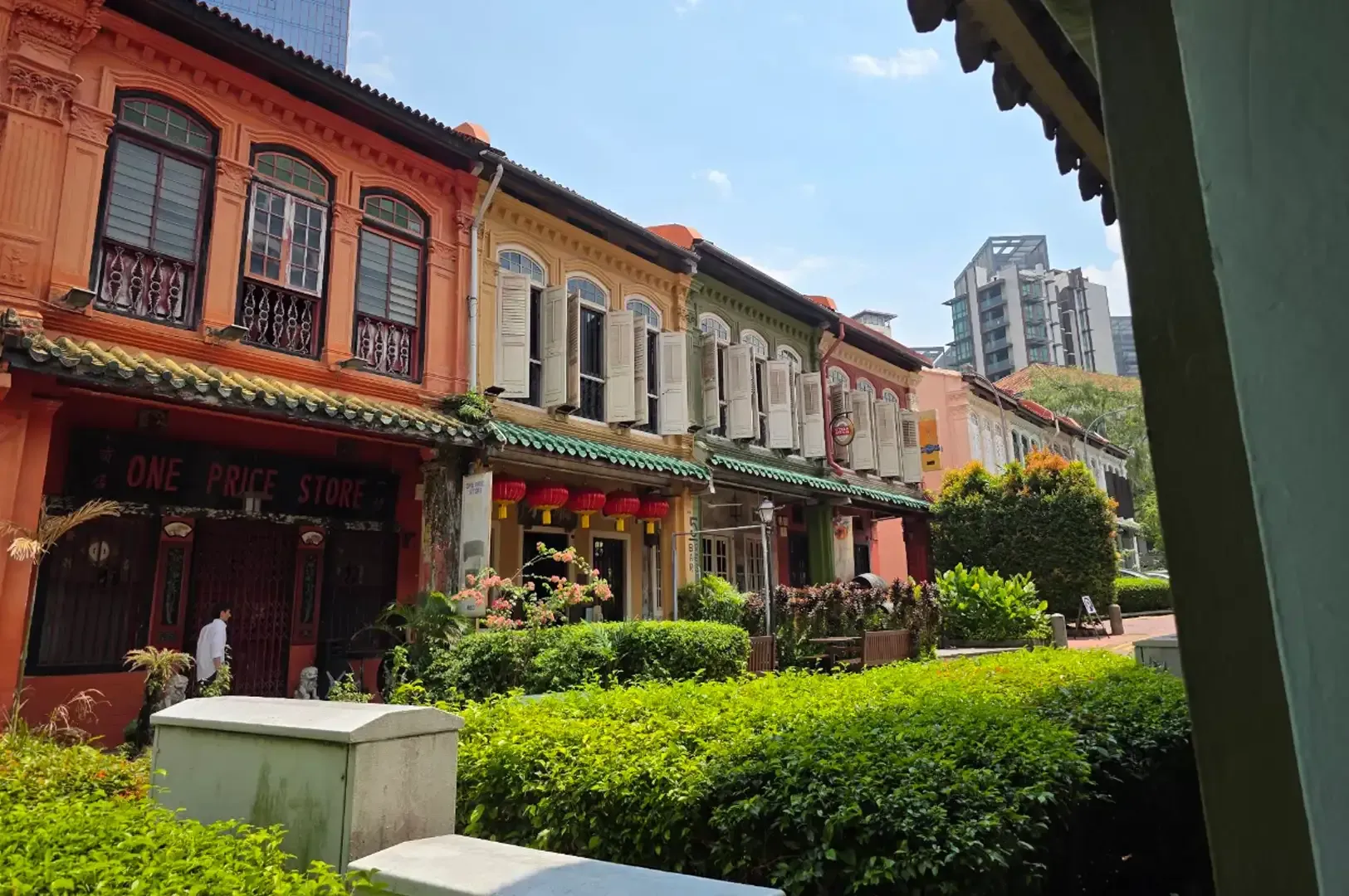 Colorful shophouses with balconies and greenery along a narrow street under a bright sky