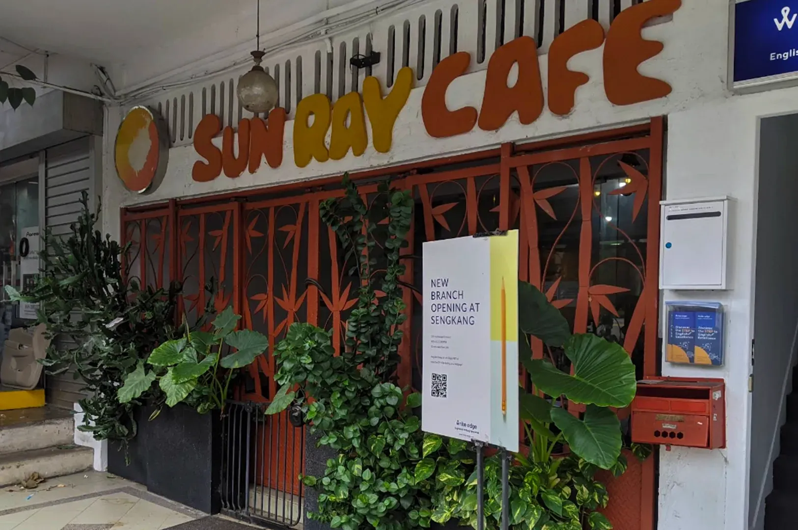 The storefront of Sun Ray Cafe, featuring yellow signage, a metal bamboo-patterned gate, and various potted green plants.
