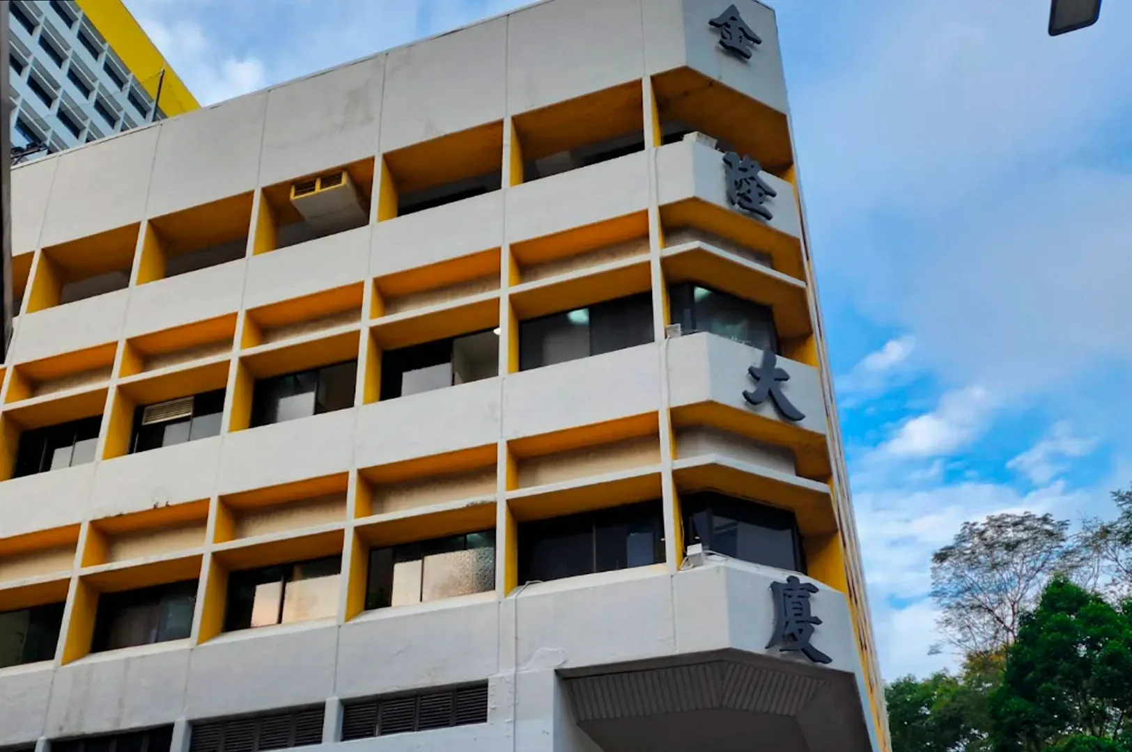 This photograph, taken from a slightly low angle, captures the exterior of a multi-story, concrete and cement building against a light blue, partly cloudy sky. The building has a modern, somewhat utilitarian style, featuring a white painted facade and recessed window panels. A striking feature is the pattern of painted bright yellow rectangular frames around most of the window and ventilation openings on the front and side facades. On the building's corner, vertically-oriented black Chinese characters are mounted, likely the building or business name. At the very top, two gold characters are visible. Green, lush tree branches and other urban elements are present in the foreground and to the side. The overall impression is an older, but painted and still active urban structure.