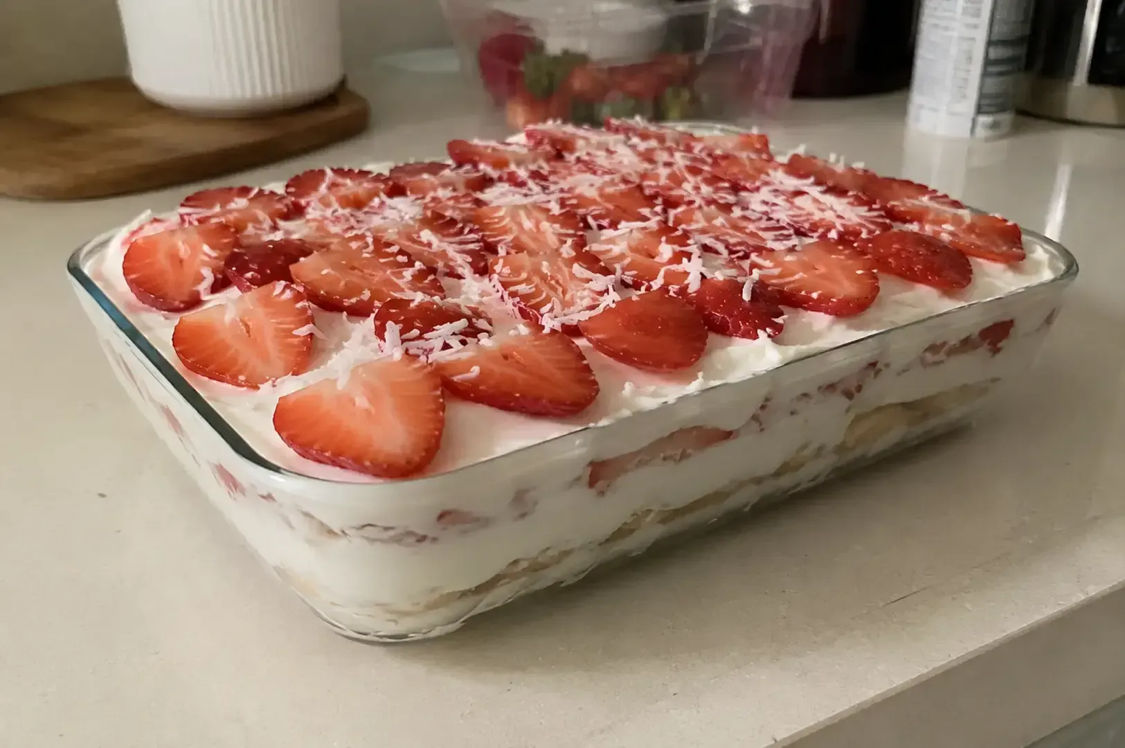 Strawberry tiramisu in a glass dish on a kitchen counter, topped with sliced strawberries and coconut flakes