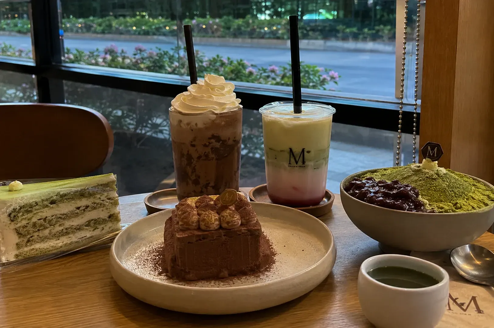 Assorted desserts and drinks on a café table by a window, with greenery outside