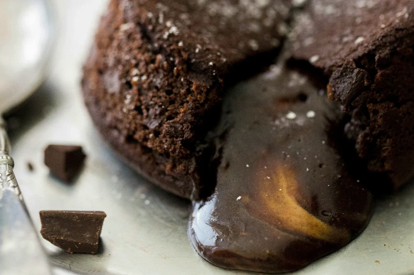 A chocolate lava cake with a warm, flowing center on a metal plate, served with a spoon and chocolate chunks.