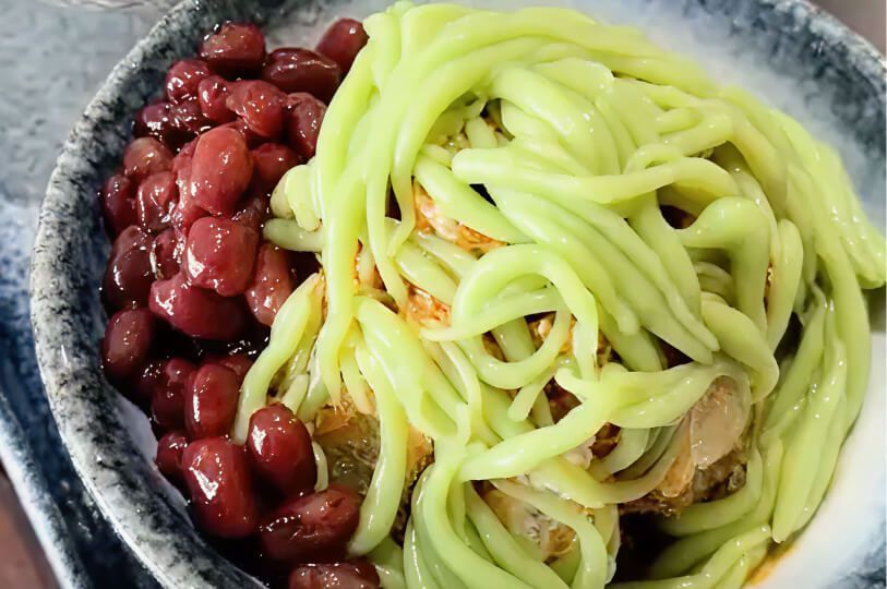 A close-up of Chendol, featuring green pandan jelly noodles and red beans over shaved ice.