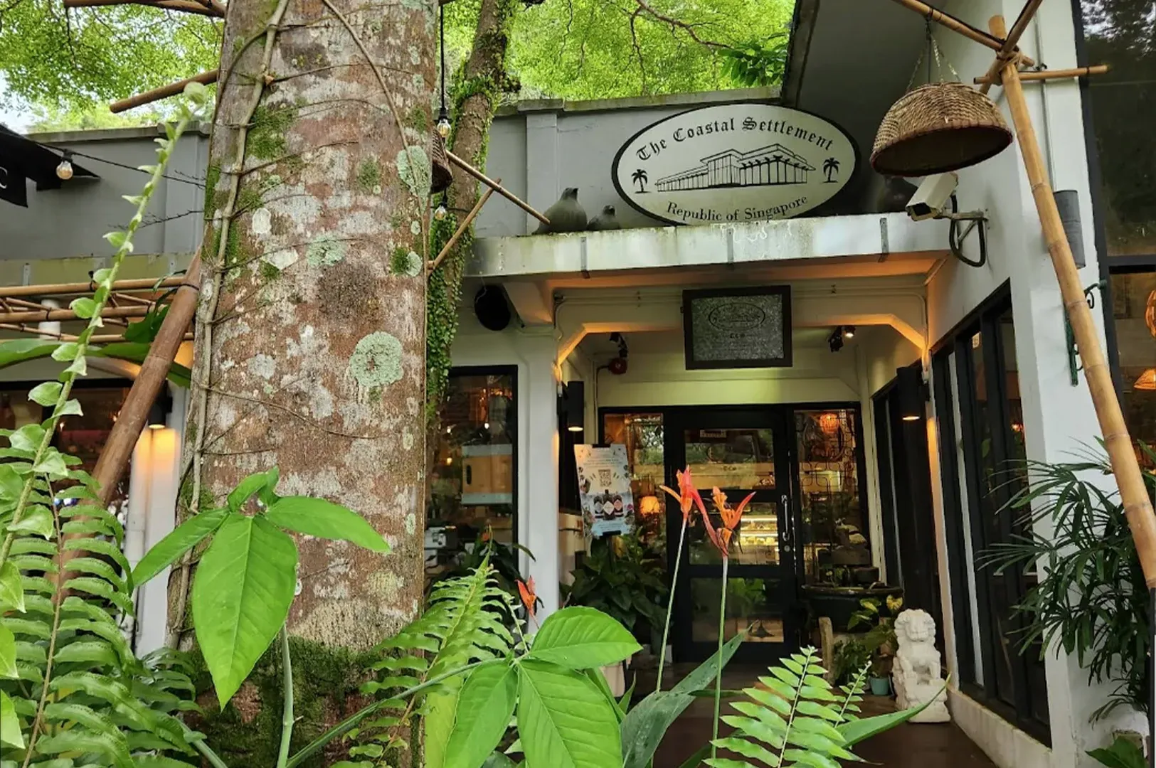 Small café entrance nestled in lush greenery, with a wooden sign and warm lights glowing inside