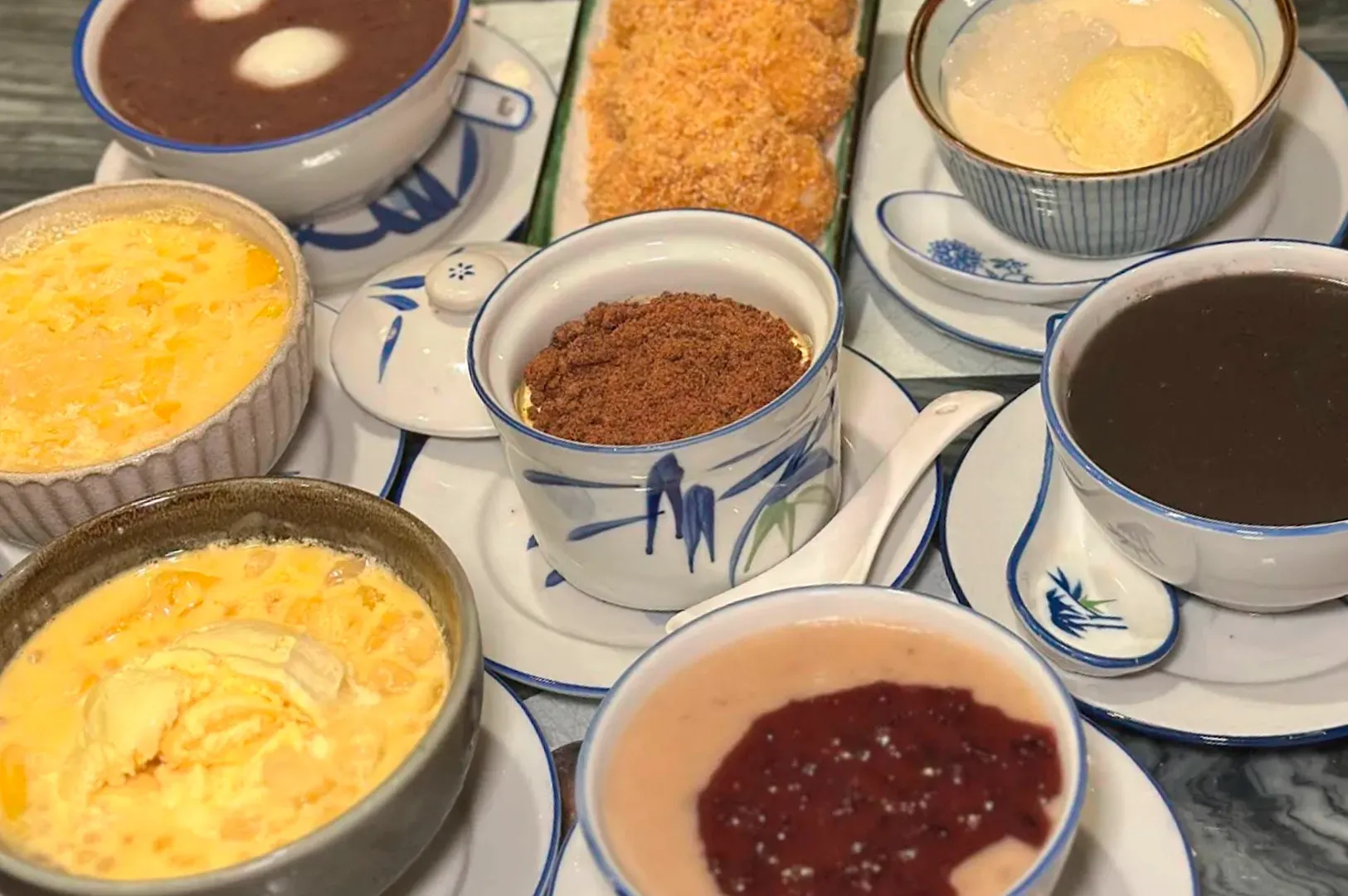 A top-down view of a table spread with a variety of traditional Chinese desserts served in classic blue-and-white porcelain bowls. Visible dishes include Mango Sago with Pomelo, Black Sesame Paste, Red Bean Soup with rice balls, and a steamed egg custard topped with red beans. The arrangement is colorful and highlights the diverse textures of the desserts.