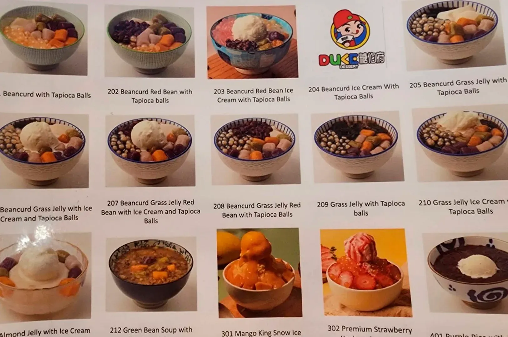 A close-up photograph of a printed menu page for Duke Dessert displays a grid of fifteen different dessert bowls, each with a corresponding number, name, and small illustrative photo. The menu features a variety of traditional and modern Asian desserts, primarily based on beancurd, grass jelly, and snow ice. Ingredients like tapioca balls, red beans, taro balls, and scoops of ice cream are visible in the colorful bowls. Notable items include 