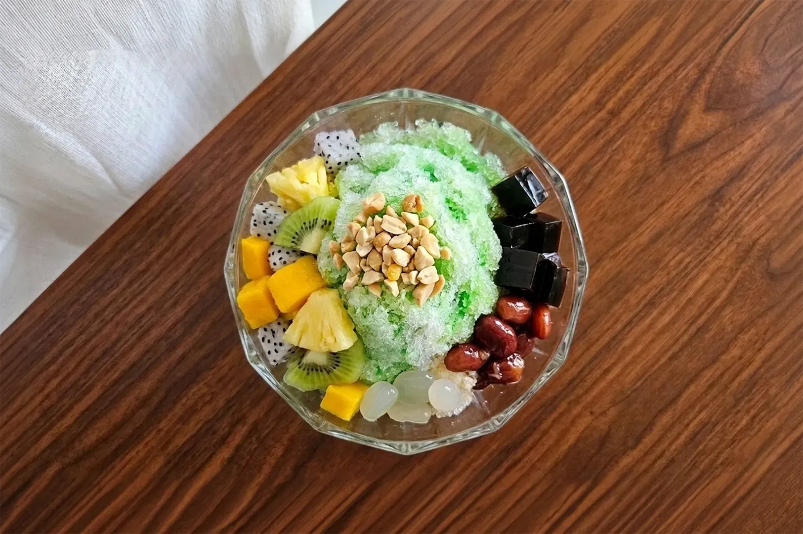 A top-down view of a clear glass bowl filled with a colorful dessert, likely shaved ice (ais kacang or bingsu). The dessert features a mound of green-tinted shaved ice topped with crushed peanuts, surrounded by colorful toppings including diced mango, dragon fruit, kiwi slices, red beans, and dark jelly cubes. The bowl sits on a polished dark wood table.