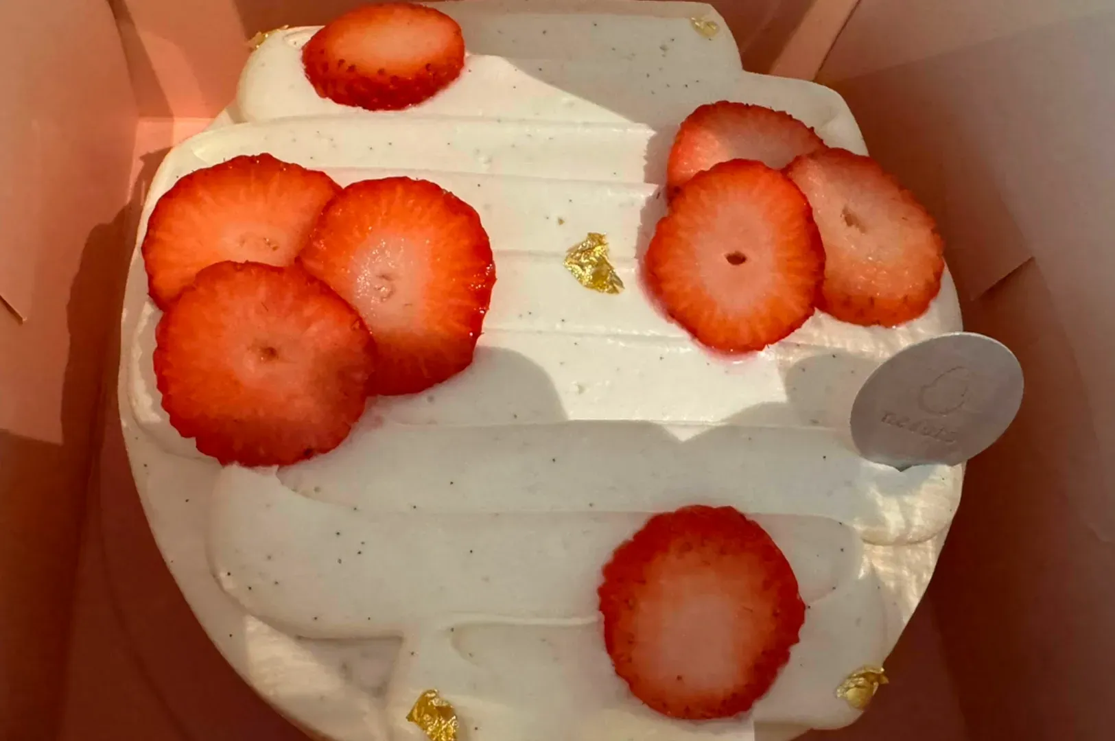 A top-down close-up of a delicate strawberry cream cake inside a soft pink box. The cake is topped with smooth, piped white cream speckled with vanilla bean. It is garnished with several thin, bright red strawberry slices and tiny flecks of edible gold leaf. A small, circular silver Nesuto brand tag is tucked into the side of the cream.