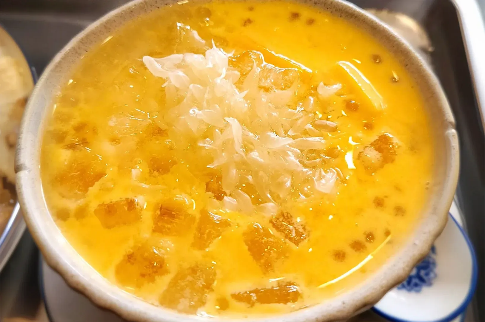 A close-up shot of a bowl of Mango Sago with Pomelo. The dessert is a bright, creamy yellow color filled with small, translucent sago pearls and chunks of fresh mango. A generous mound of pale, shredded pomelo pulp sits in the center, glistening under the light.
