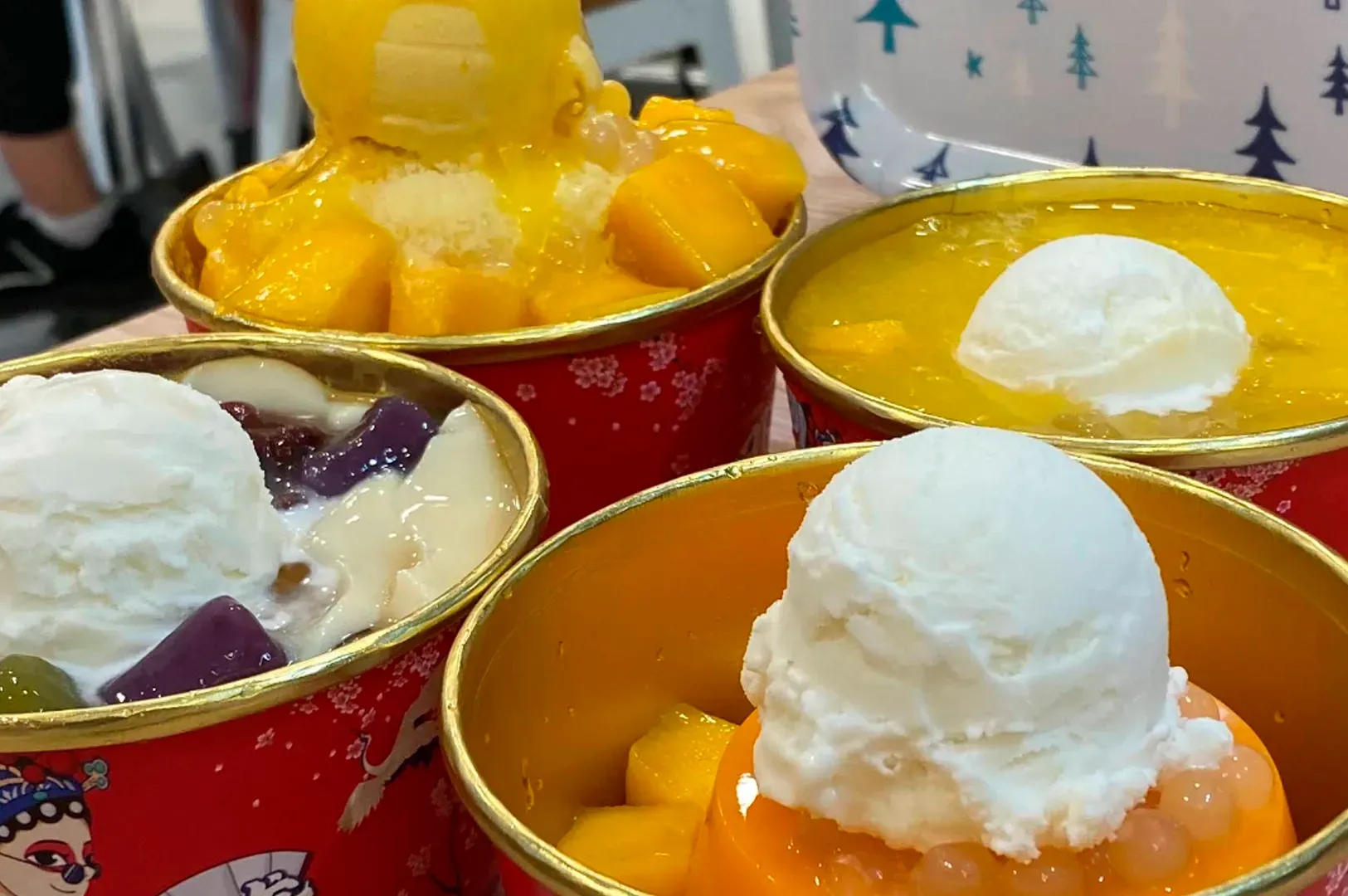 A high-angle, close-up photograph features four vibrant dessert bowls from Duke Dessert arranged closely together on a table. Each bowl is red with a gold rim and festive Chinese-style illustrations. The top-left bowl is piled high with bright yellow mango chunks, mango sauce, and a scoop of yellow ice cream. The top-right bowl contains a clear jelly topped with a large scoop of white ice cream. The bottom-left bowl features silky white beancurd, purple taro balls, red beans, and a scoop of white ice cream. The bottom-right bowl shows a bright orange mango pudding topped with a large scoop of white ice cream and translucent pink pearls. The lighting is bright and flat, highlighting the textures of the various toppings.
