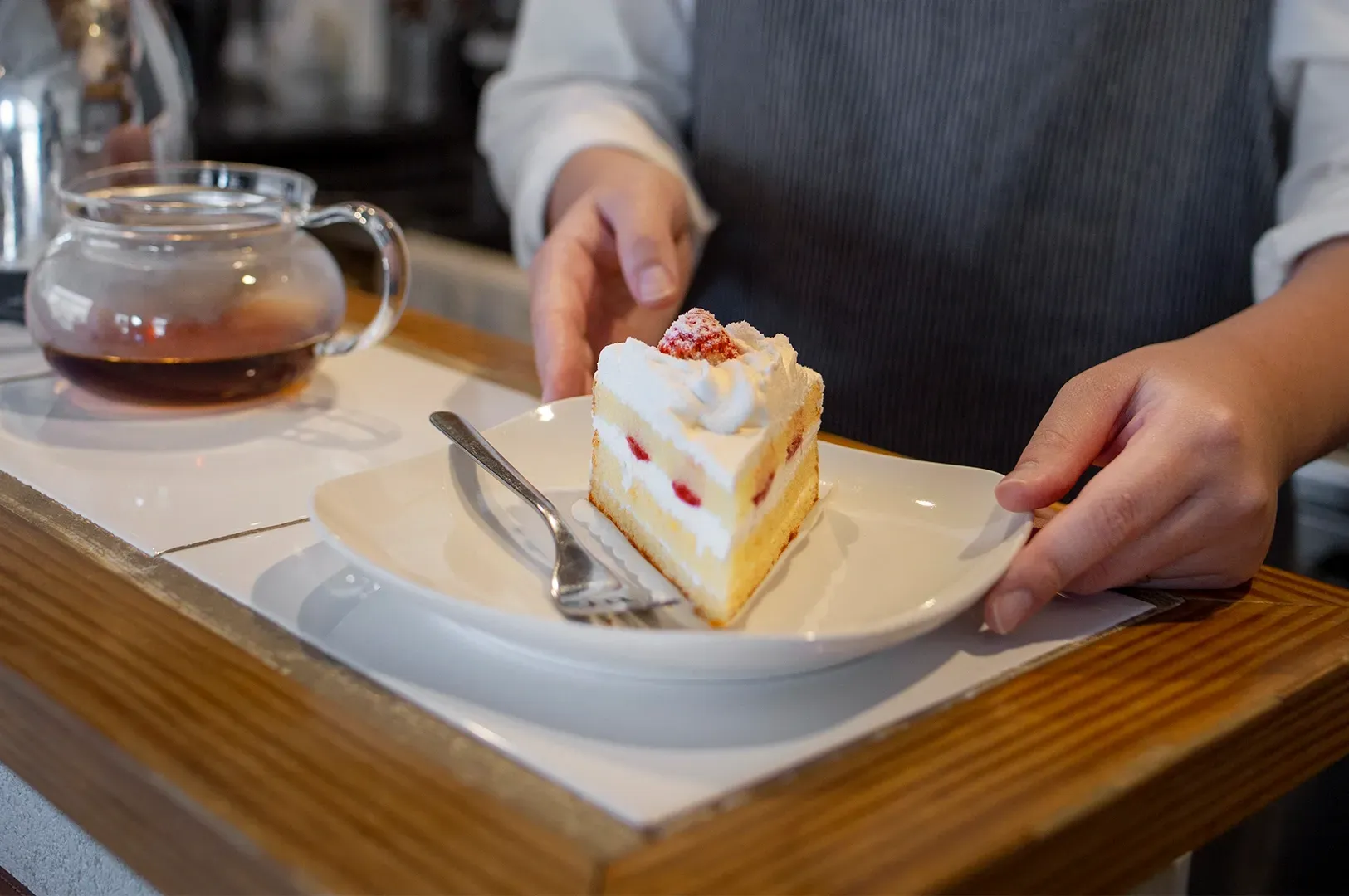 A person in a grey apron holds a white plate featuring a single slice of strawberry cream cake. The cake has light sponge layers with whipped cream and sliced strawberries, topped with half a fresh strawberry. A glass teapot filled with dark tea sits in the blurred background on a wooden counter.