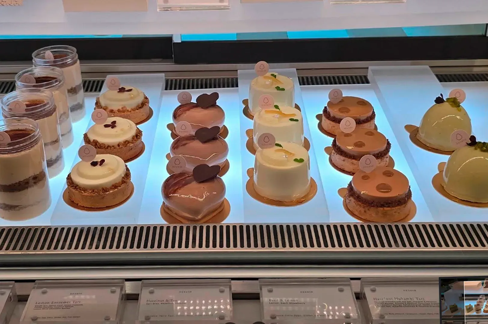 A brightly lit refrigerated display case filled with rows of meticulously crafted French-style entremets and tarts. The selection includes jars of tiramisu, white lemon tarts, heart-shaped chocolate mousses, and glossy pistachio domes. Each pastry is uniform in shape and topped with a signature round Nesuto logo tag. Small descriptive labels are visible at the bottom of the glass case.