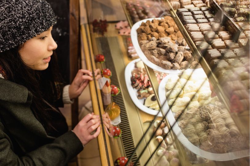 A young woman in a beanie looking through a glass display case filled with various gourmet chocolates.