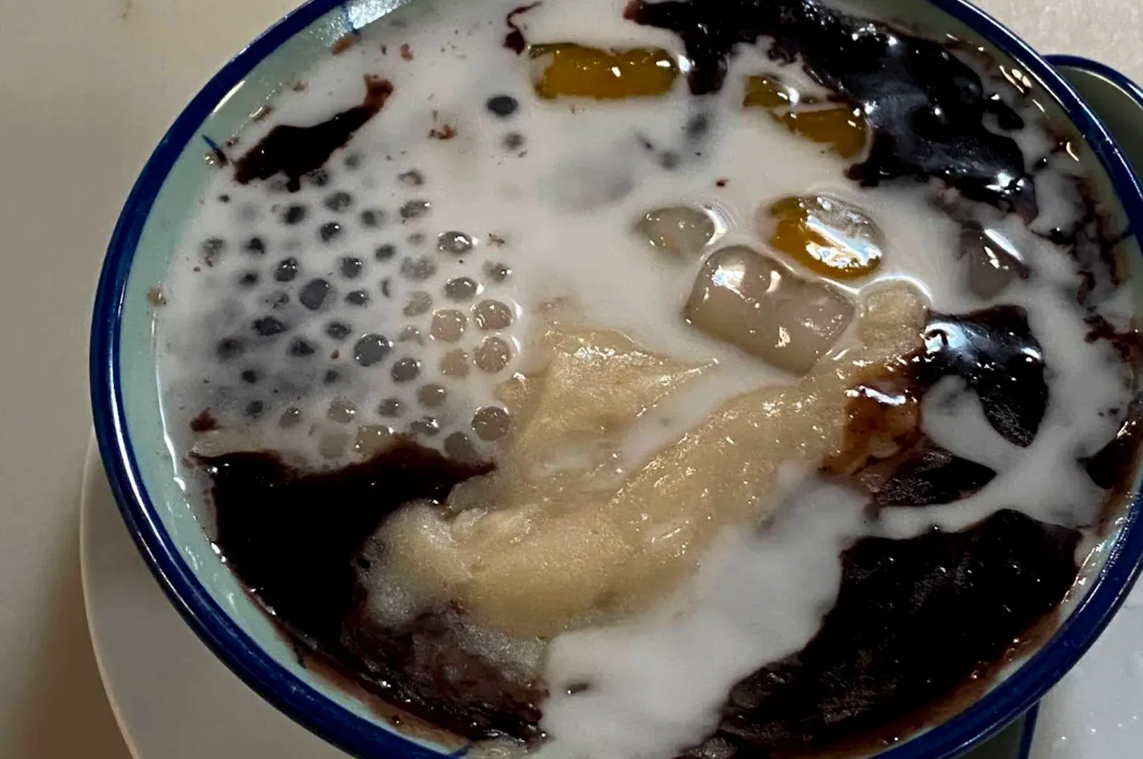 A bowl of Pulut Hitam (black glutinous rice porridge). The deep purple, almost black rice is partially swirled with a thick, marble-like drizzle of coconut milk. Floating within the mixture are translucent, chewy attap seeds and golden sweet potato cubes, adding layers of texture and natural sweetness to the traditional warm dessert.