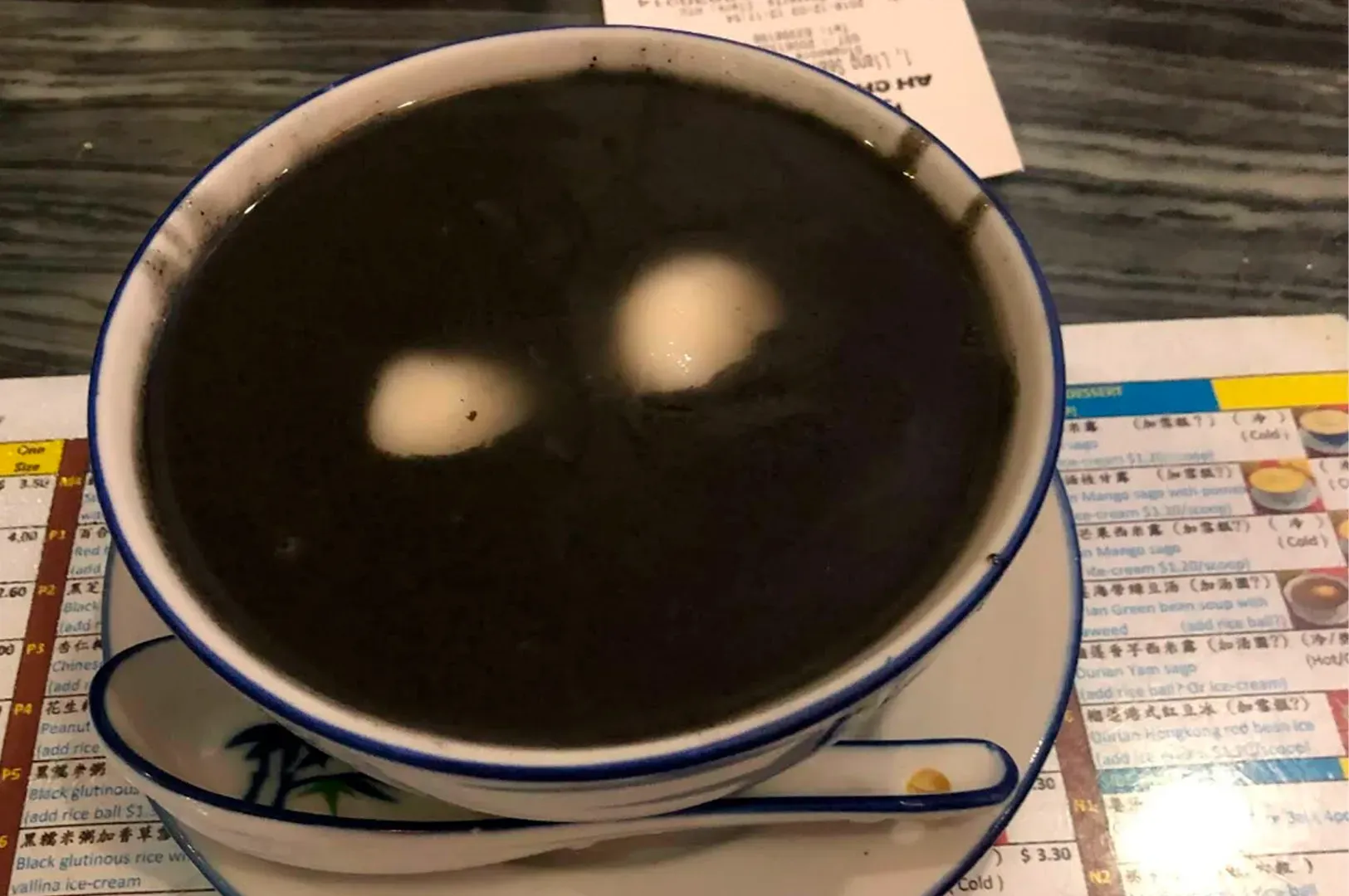 A close-up of a bowl of warm Black Sesame Paste, a thick, glossy, jet-black dessert soup. Two round, white glutinous rice balls (tang yuan) are partially submerged in the center, creating a sharp color contrast. The bowl is set on a matching saucer with a traditional Chinese spoon resting beside it.