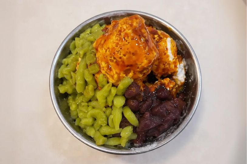 A metal bowl filled with Chendol, featuring green rice flour jelly, red beans, and shaved ice topped with a large scoop of durian flesh and drizzled with gula melaka syrup.