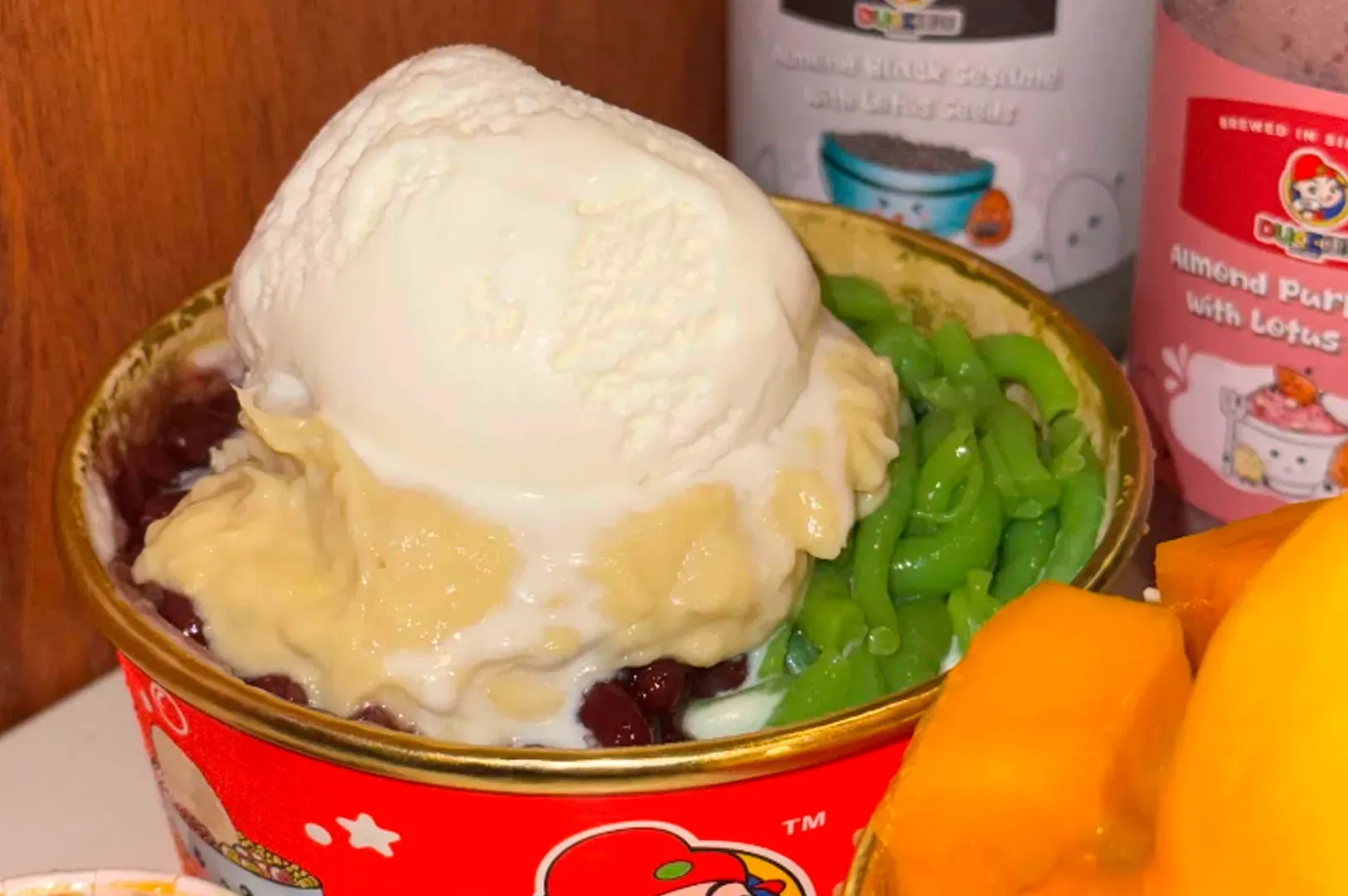 A macro, top-down photograph captures a detailed view of a Durian Chendol dessert in a red and gold paper bowl. A large, prominent scoop of white coconut ice cream sits atop a thick, pale yellow dollop of creamy durian flesh. Surrounding the durian are vibrant green pandan jelly noodles (chendol) and a bed of dark, glossy red beans. The bowl is positioned in the foreground, with part of another dessert and two bottled drinks from the same shop visible in the soft-focus background. The image has a warm, slightly yellow tint, emphasizing the rich colors of the dessert ingredients.