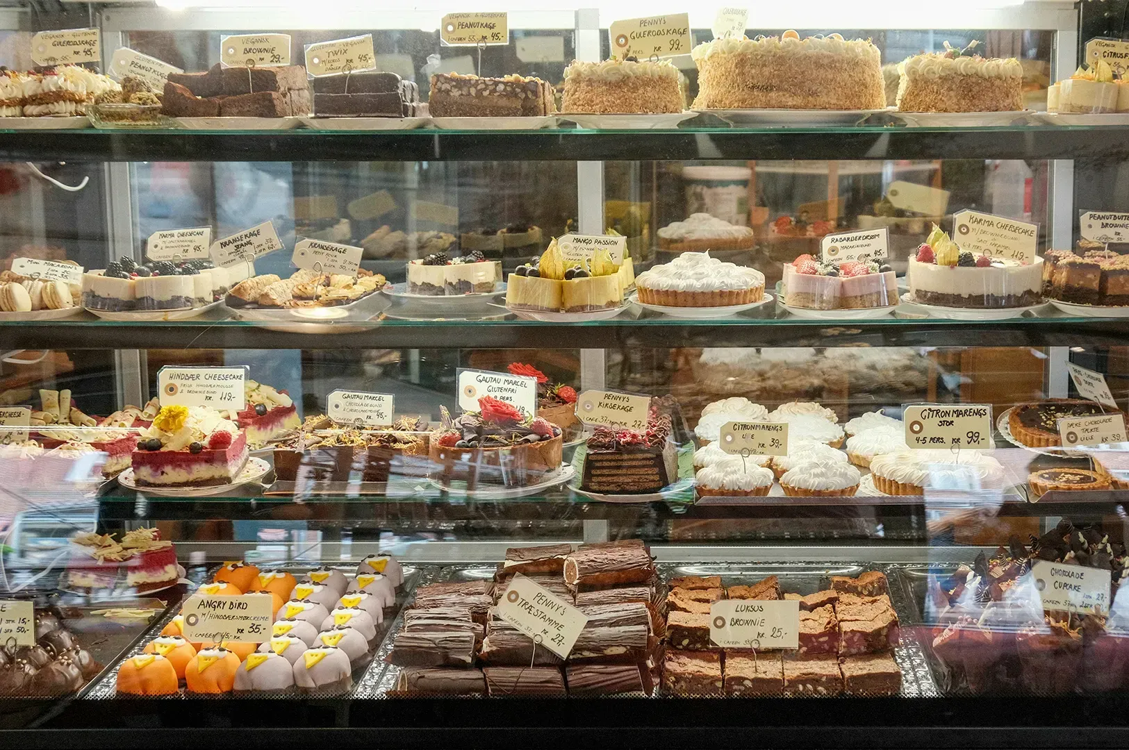 A view through a glass display case at a bakery, showing several shelves stocked with a wide variety of desserts. The selection includes cheesecakes with fruit toppings, chocolate brownies, tarts, and uniquely decorated treats, all labeled with small price tags.