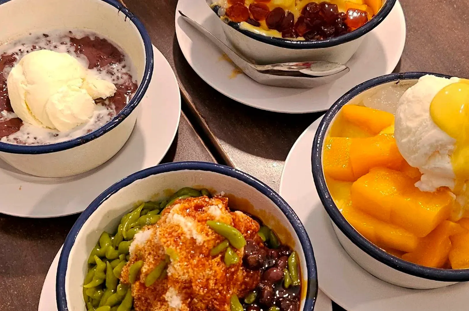 A vibrant assortment of four different desserts is presented here in white enamel-style bowls with blue rims. One bowl features mango pomelo sago topped with a large scoop of vanilla ice cream and mango puree. Another contains a chilled grass jelly base loaded with colorful taro balls and pearls. The bottom two bowls showcase the Chendol with thick gula melaka syrup and a warm red bean soup paired with a melting scoop of ice cream, illustrating the shop's blend of hot and cold offerings.