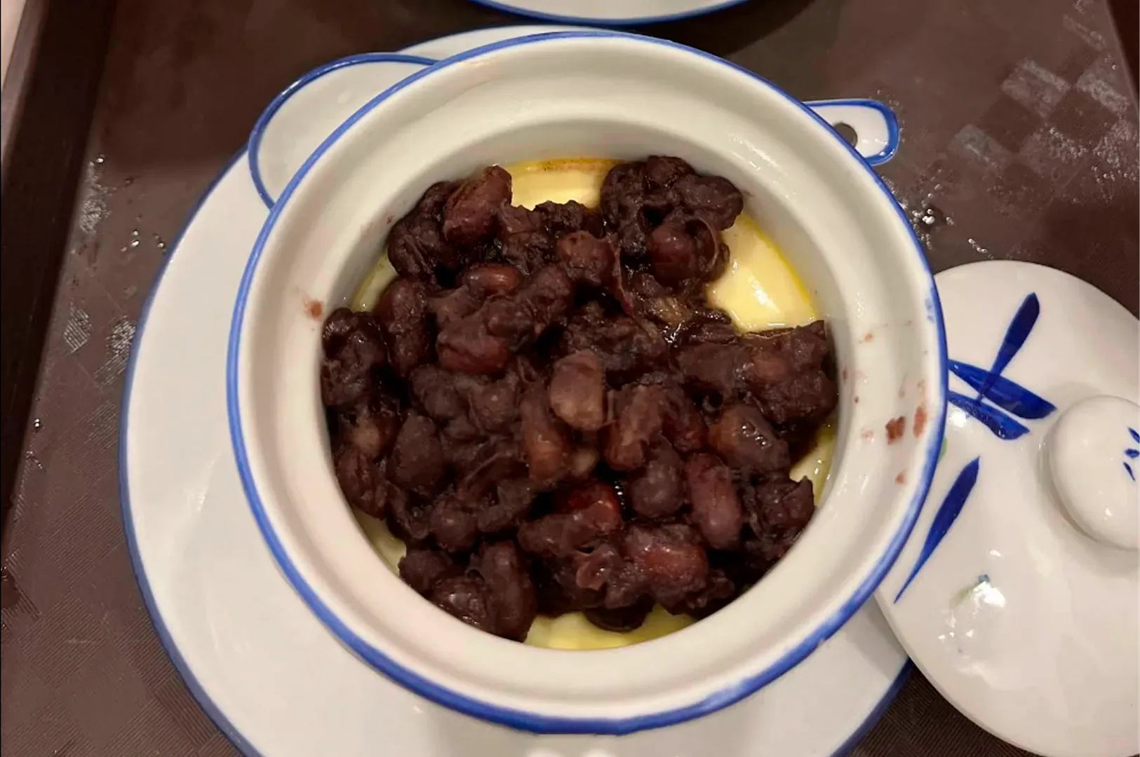 A bird's-eye view of a steamed milk custard served in a small porcelain pot. The smooth, pale yellow custard is topped with a dense layer of soft, sweetened whole red beans. The pot's lid, featuring a blue bamboo leaf pattern, sits to the side on a dark brown tray.