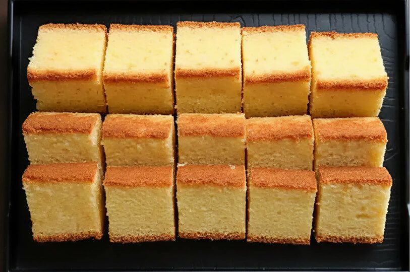 A black tray neatly filled with two rows of rectangular slices of light yellow sponge cake.