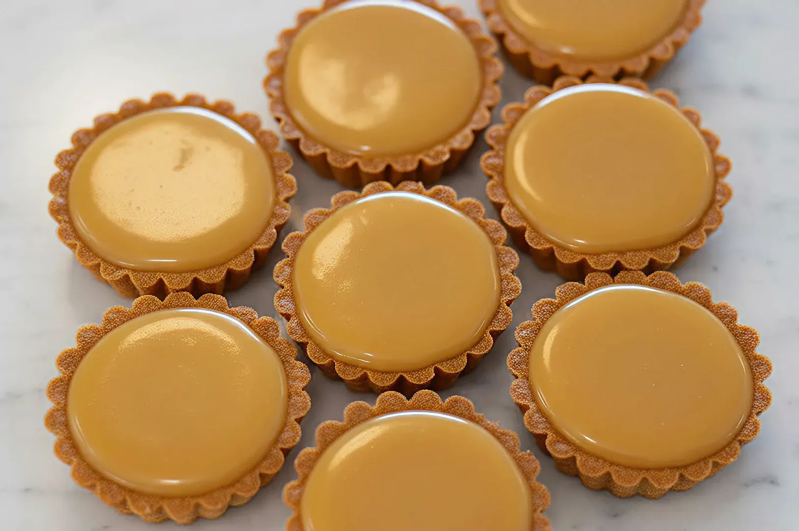 A top-down view of seven small, round tarts arranged on a white marble surface. Each tart has a fluted crust and is filled with a smooth, glossy, caramel-colored custard.