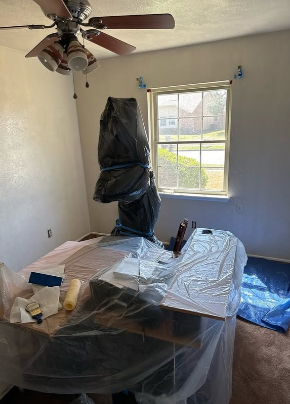 Room interior with furniture covered in plastic for painting. Window and ceiling fan visible.