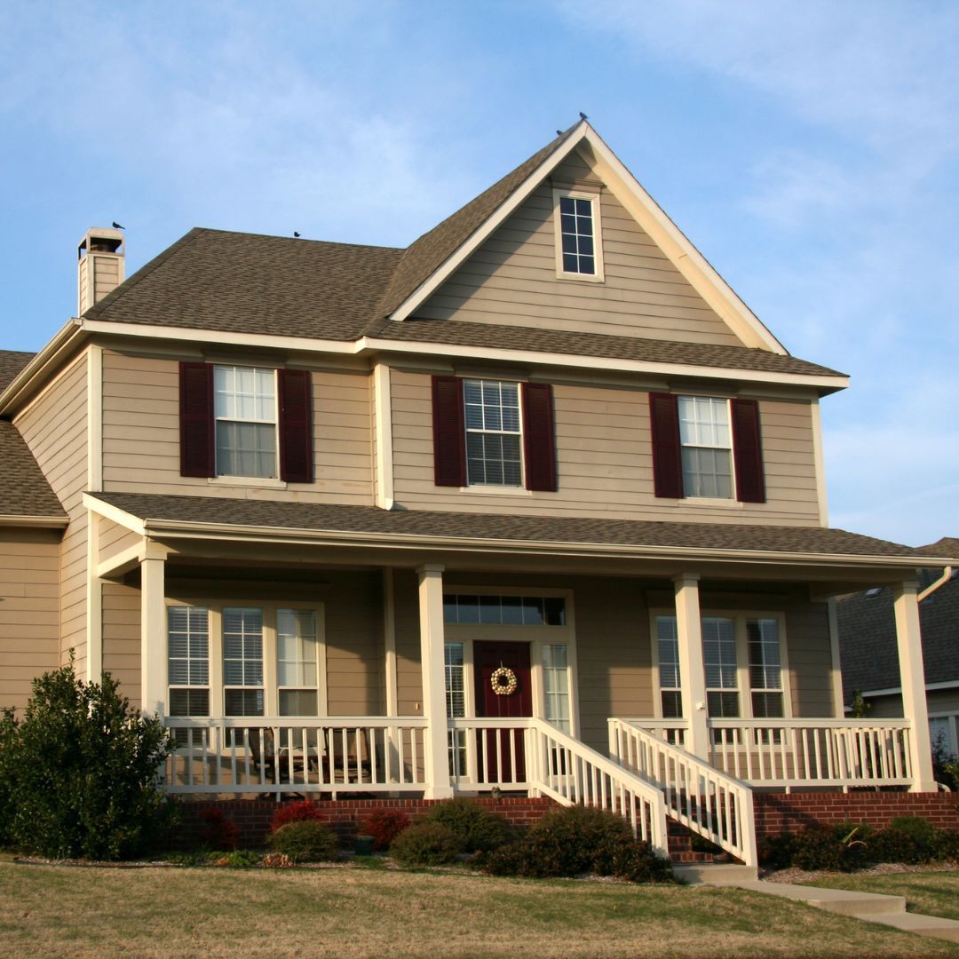 Two-story beige house with a porch and red shutters, under a clear blue sky.