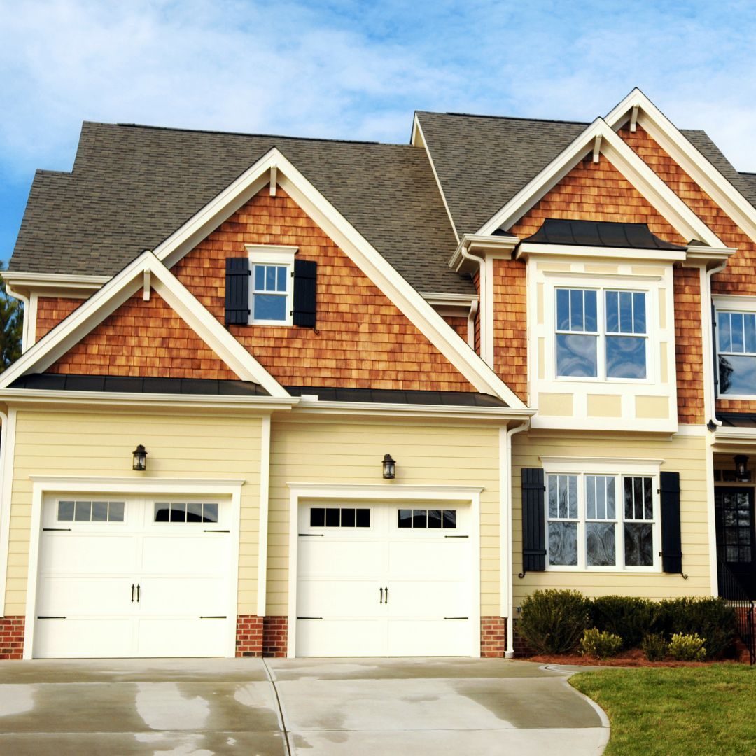Two-story house with tan siding, brown cedar shake accents, white garage doors, and blue sky.