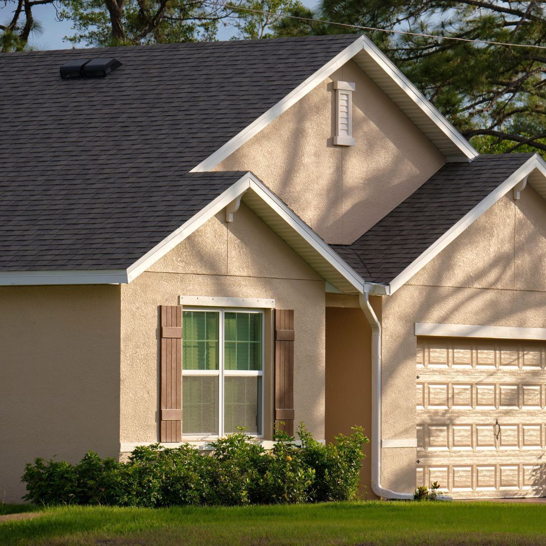 Tan house with dark roof and brown shutters, green lawn, trees in background.