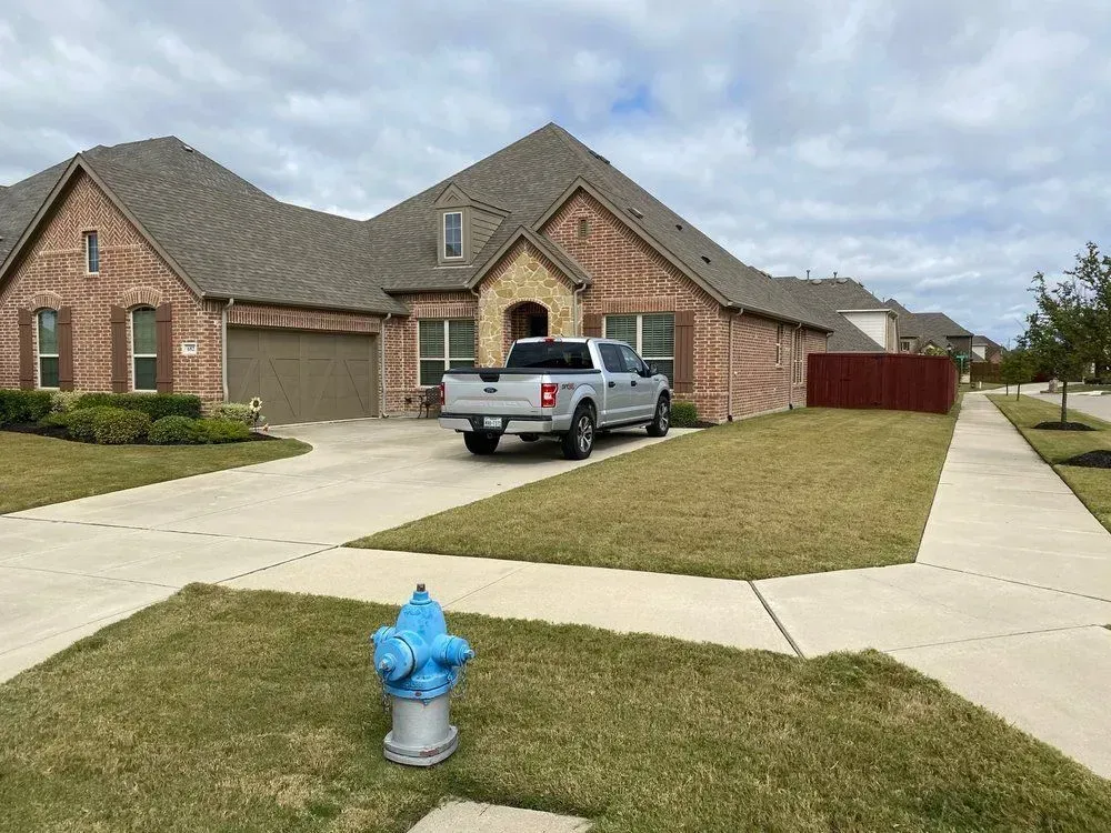 A silver pickup truck parked in the driveway of a brick house with brown roof; blue fire hydrant in foreground.