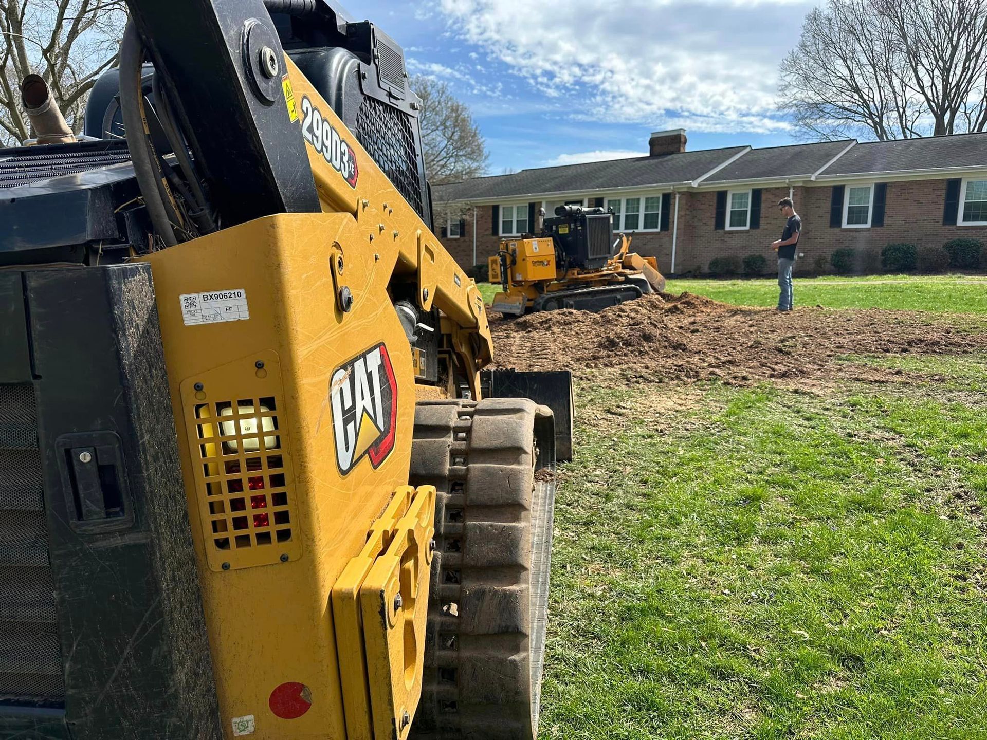 A cat bulldozer is sitting in a grassy field in front of a house.