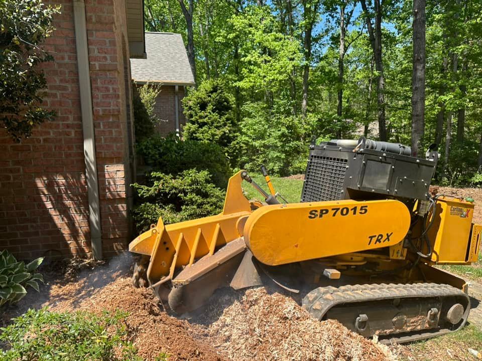 A yellow tractor is cutting a tree stump in front of a brick building.