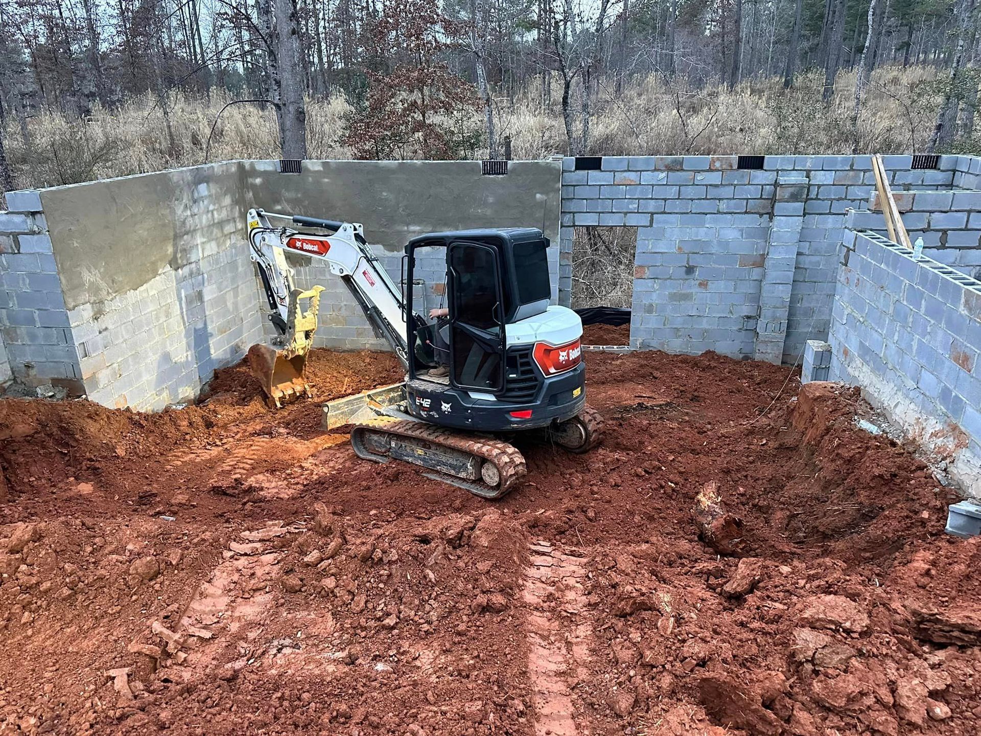 A small excavator is digging a hole in the dirt in front of a brick wall.