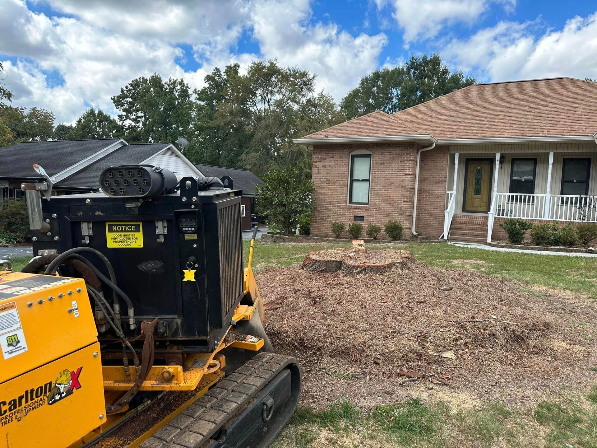 A yellow tractor is working on a tree stump in front of a brick house.