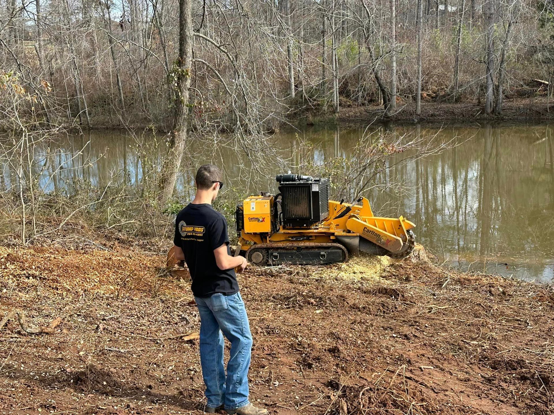 A man is standing in front of a stump grinder next to a river.