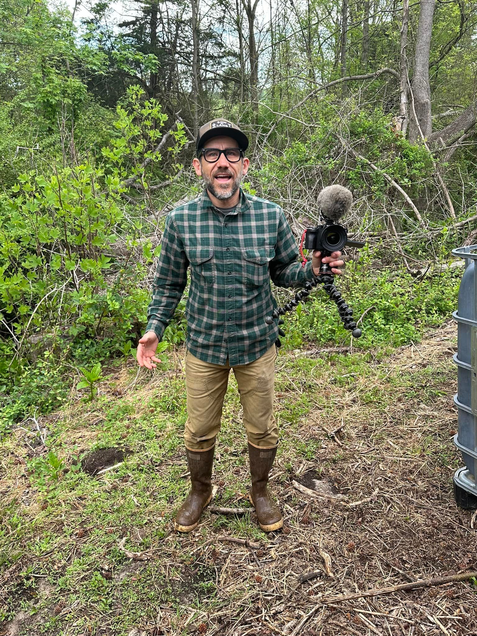 A man in a plaid shirt is standing in the woods holding a microphone.
