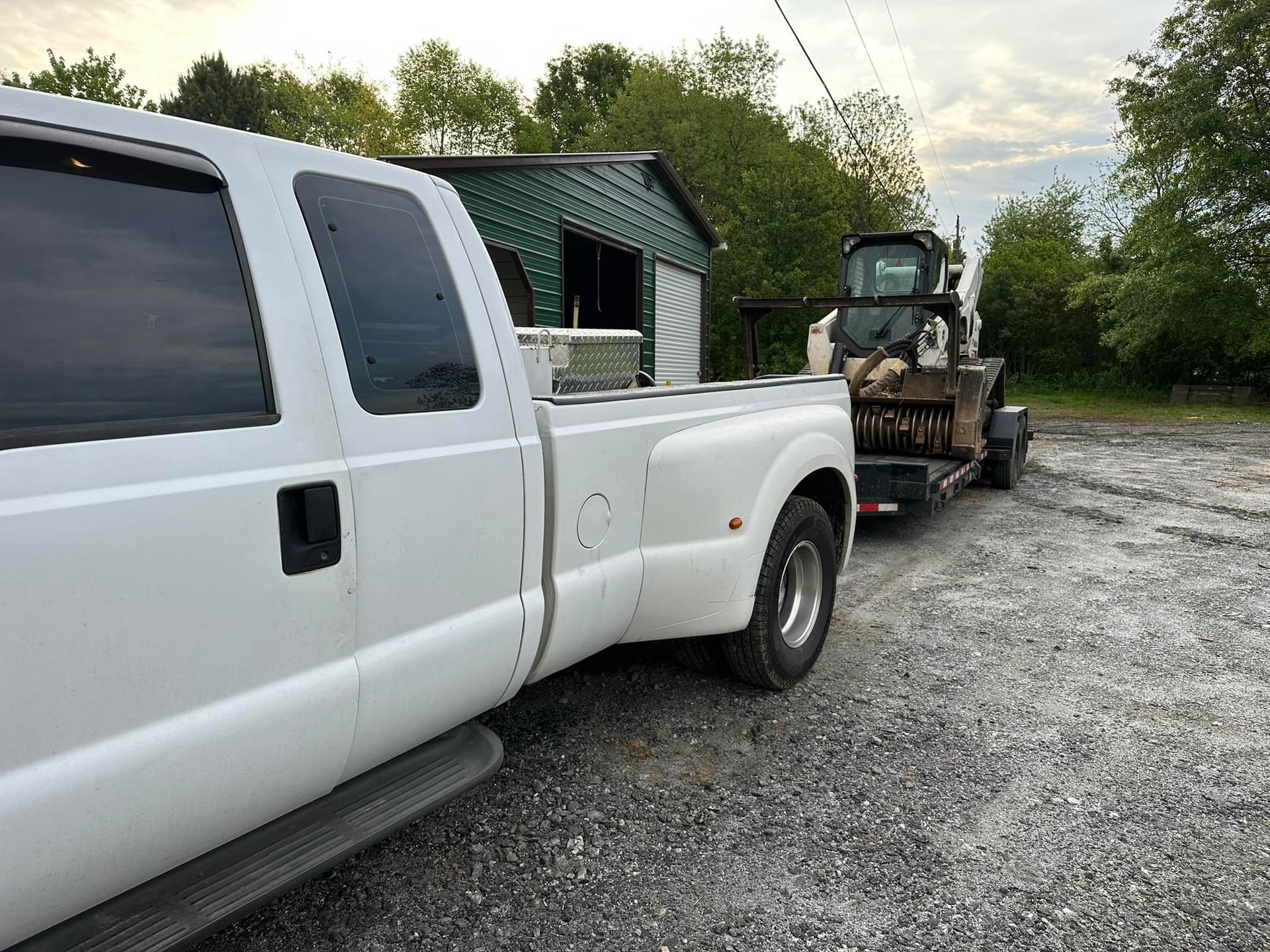 A white truck is towing a tractor in a gravel lot.
