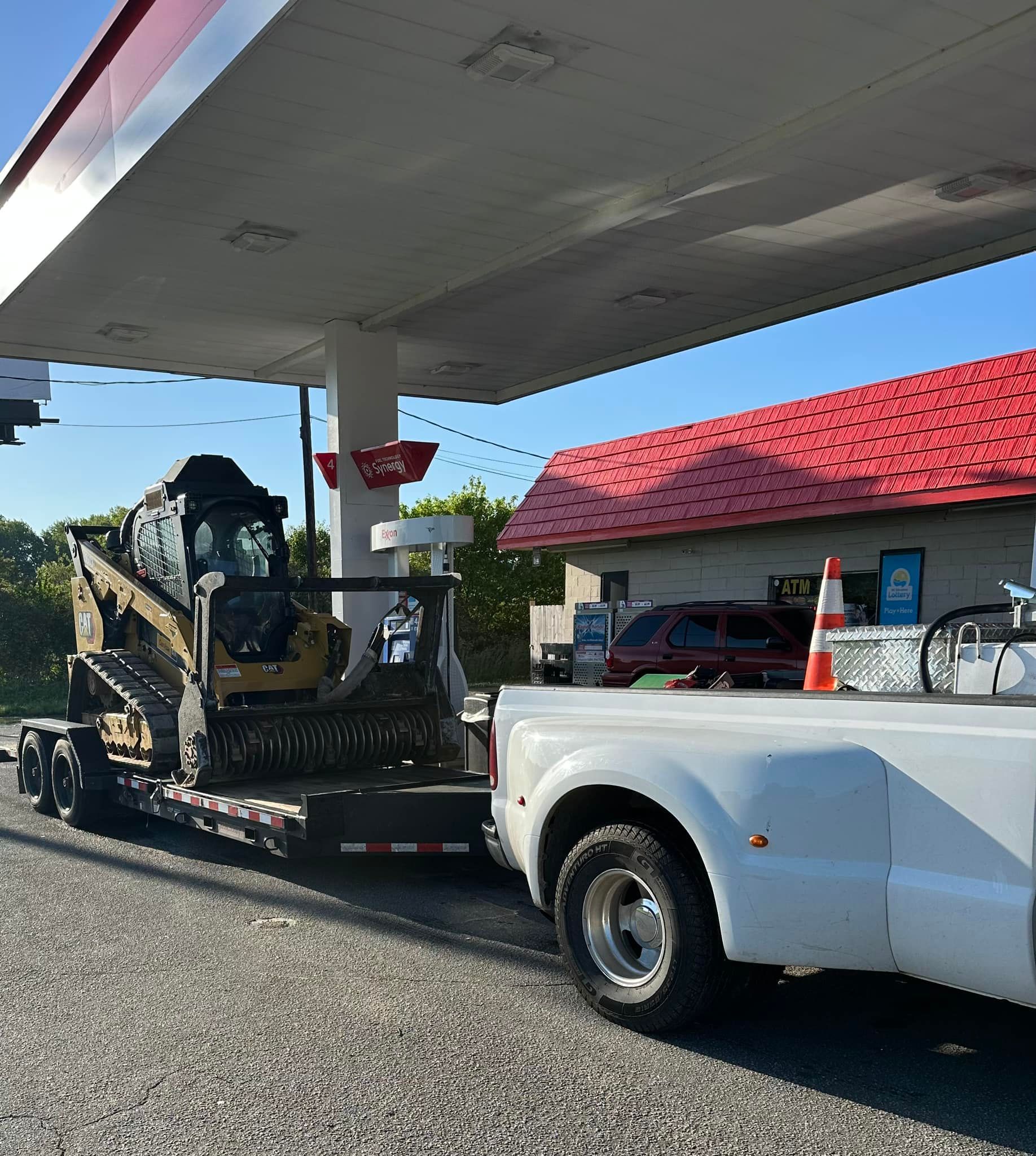 A white truck is towing a bulldozer at a gas station.